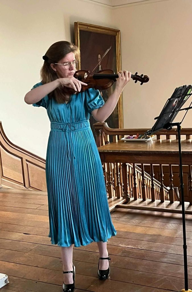 Une femme jouant du violon, vêtue d'une robe plissée bleu turquoise, avec des chaussures à talons, dans une pièce au décor classique.