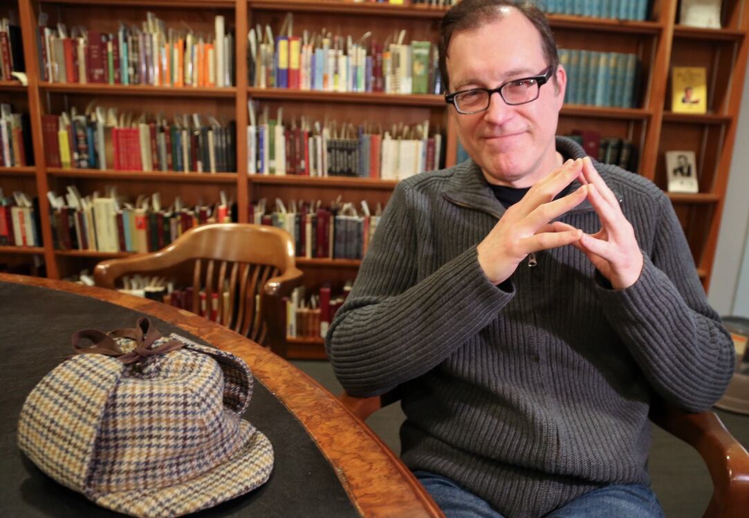 Un homme souriant assis à une table avec une casquette à carreaux de style Sherlock Holmes, entouré de livres dans une bibliothèque.