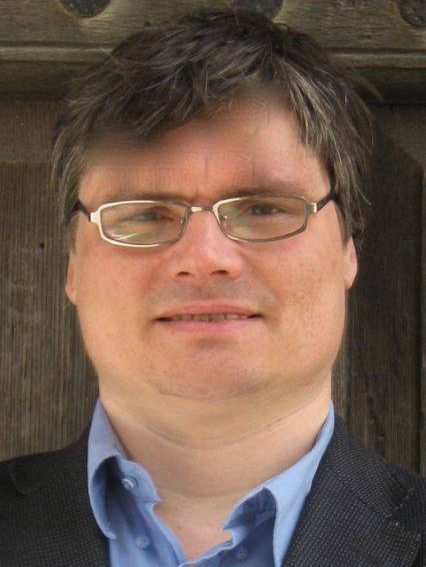 Portrait d'un homme avec des cheveux bruns et des lunettes, portant une chemise bleu clair et un blazer noir, devant un fond en bois.